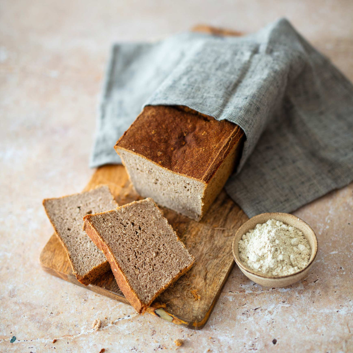 Ein angeschnittenes, saftiges Brot aus der Pfundskerl-Brotbackmischung liegt auf einem Holzbrett, teils mit einem grauen Leinentuch bedeckt. Zwei Brotscheiben sind vorn aufgefächert. Rechts daneben steht eine kleine Schale mit Mehl.