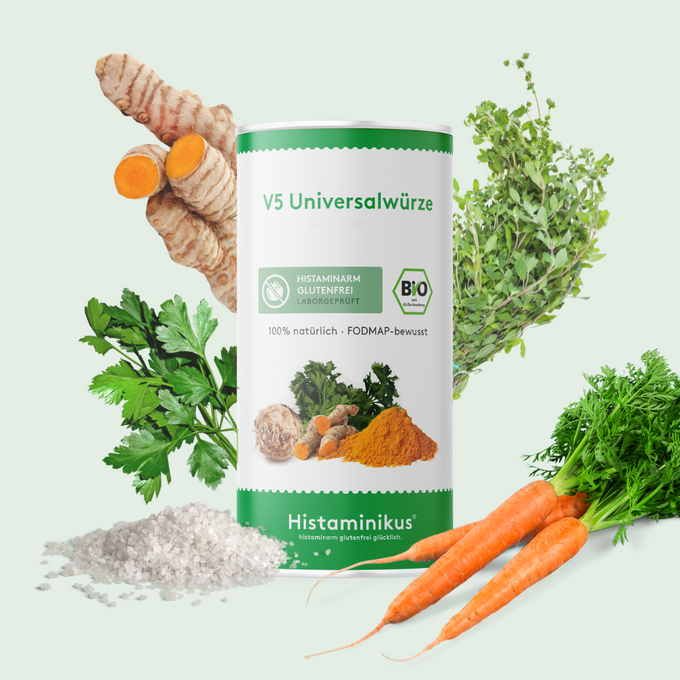 Dose der Histaminikus V5 Universalwürze vor hellem Hintergrund, umgeben von einzelnen Zutaten: frische Kurkumawurzel, Majoran, Petersilie, Karotten und grobes Steinsalz.