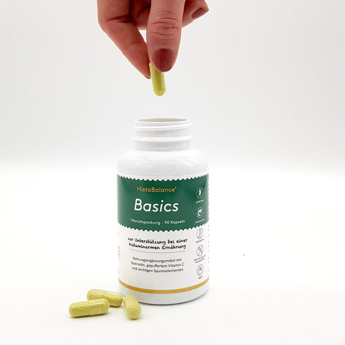 Weiße Dose mit grünem Etikett des Produkts „HistaBalance Basics“, offenstehend auf weißem Hintergrund. Eine Hand hält eine gelbe Kapsel über die Dose. Vor der Dose liegen drei weitere gelbe Kapseln auf dem Tisch. Etikett zeigt Aufschrift zur Unterstützung bei histaminarmer Ernährung.