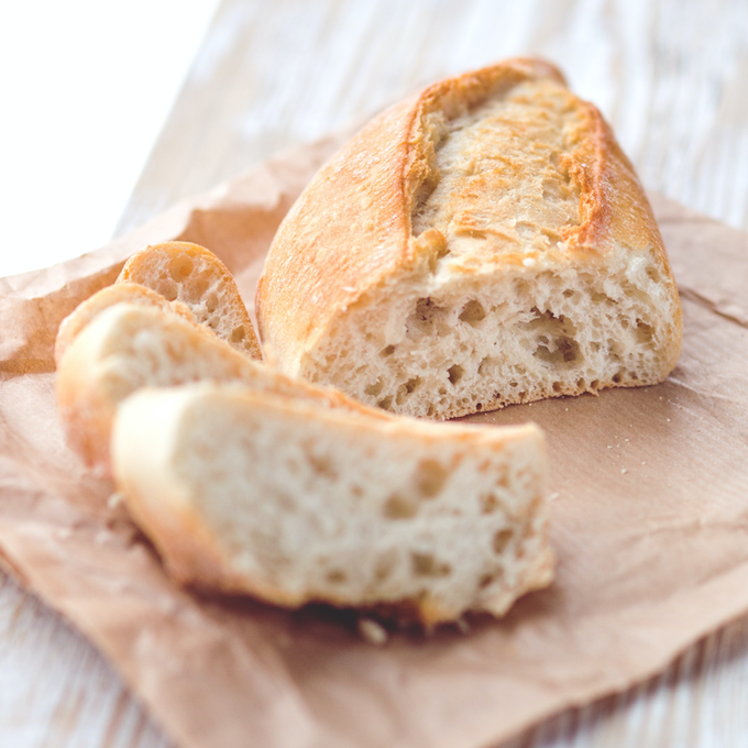 Nahaufnahme eines frisch gebackenen Baguettes auf braunem Papier: Im Vordergrund zwei angeschnittene Scheiben mit luftiger Krume, dahinter ein goldbraun gebackenes Baguettestück mit knuspriger Kruste. Die helle Holzoberfläche und das weiche Licht unterstreichen den natürlichen, handgemachten Charakter.
