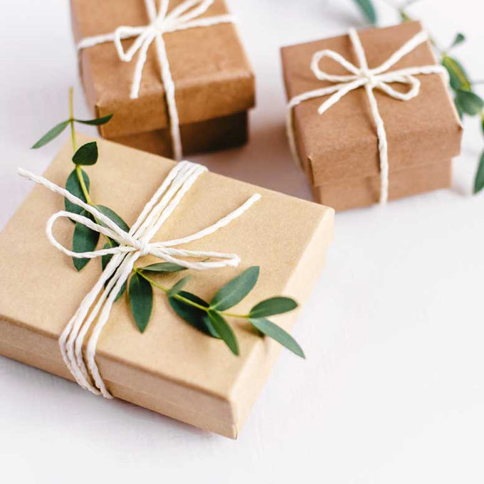 Drei in braunes Kraftpapier eingepackte Geschenke, dekoriert mit weißer Kordel und frischen grünen Zweigen, auf hellem Untergrund arrangiert.