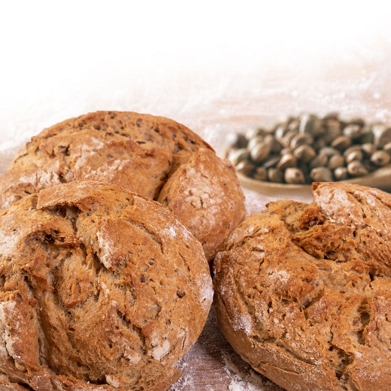 Drei runde, rustikal gebackene Brötchen mit bemehlter Kruste auf einer bemehlten Holzoberfläche. Im Hintergrund ein kleiner Teller mit Hanfsamen.