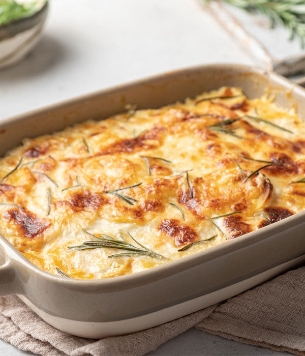 Frisch gebackenes Kartoffelgratin in einer beigen Auflaufform, goldbraun überbacken mit Rosmarinzweigen garniert, serviert auf einem hellen Stofftuch.