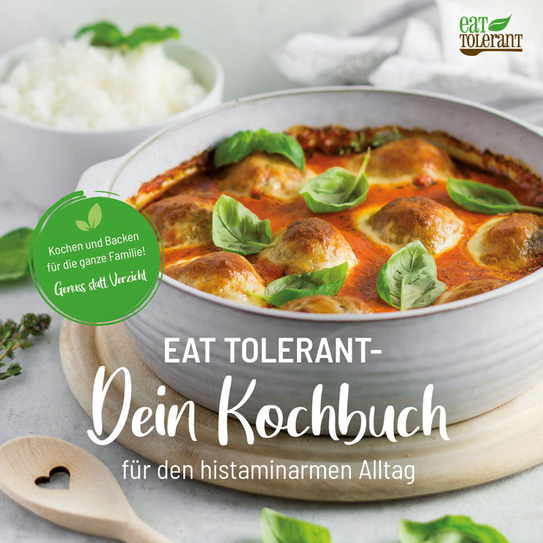 Foto eines histaminarmen Kochbuchcovers der Marke „Eat Tolerant“. Im Mittelpunkt eine weiße Auflaufform mit gebackenen Bällchen in roter Soße, garniert mit frischem Basilikum. Links ein grüner Kreis mit dem Text „Kochen und Backen für die ganze Familie! Genuss statt Verzicht“. Unten die Schrift „EAT TOLERANT – Dein Kochbuch für den histaminarmen Alltag“ in Weiß auf grauem Untergrund. Im Hintergrund ein Schälchen mit Reis und ein Holzlöffel mit ausgestanztem Herz.