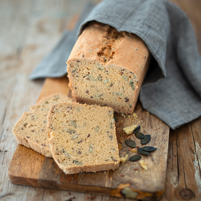 Aufgeschnittenes Körnerbrot auf einem Holzbrett. Im Anschnitt gut sichtbare Sonnenblumenkerne, Kürbiskerne und Leinsamen. Zwei Scheiben liegen im Vordergrund, daneben lose Kerne und Krümel. Das Brot ist mit einem grauen Tuch teilweise abgedeckt. Rustikaler Holztisch als Untergrund.