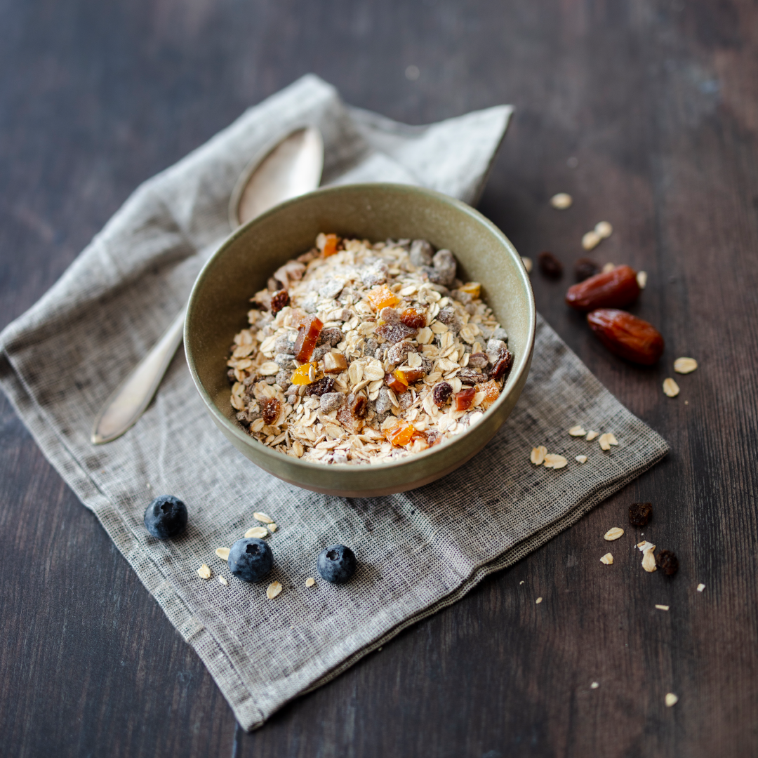 Schale mit Müslimischung auf einem grauen Leinentuch, darunter dunkler Holztisch. Im Müsli sichtbare Haferflocken, Trockenfrüchte und Kerne. Daneben liegen Datteln, Rosinen und einige Blaubeeren. Ein Löffel liegt links neben der Schale.