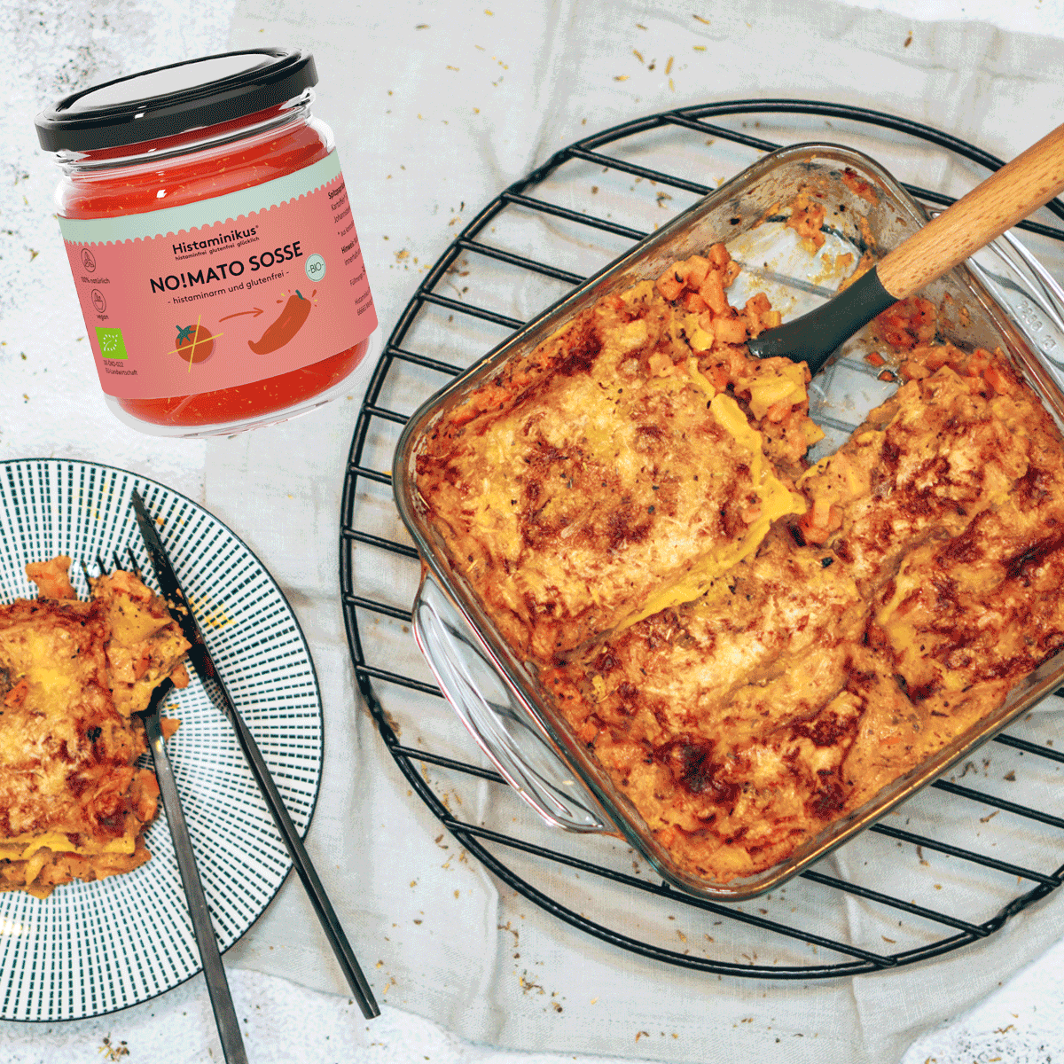 Rechts eine Auflaufform mit Lasagne auf einem schwarzen Kuchengitter. Am linken Bildrand ein schwarz-weißer Teller mit Lasagne. In der oberen linken Bildecke ist ein Glas der No!Mato Sosse von Histaminikus zu sehen.