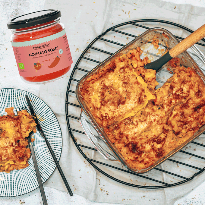Rechts eine Auflaufform mit Lasagne auf einem schwarzen Kuchengitter. Am linken Bildrand ein schwarz-weißer Teller mit Lasagne. In der oberen linken Bildecke ist ein Glas der No!Mato Sosse von Histaminikus zu sehen.