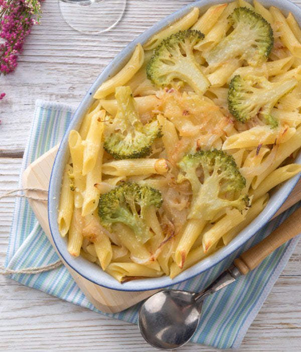 Auflaufform mit überbackenem Nudel-Gemüse-Gratin aus Penne, Brokkoli und geschmolzenem Käse, serviert auf einem Holzbrett mit gestreifter Serviette und Löffel auf hellem Holztisch.