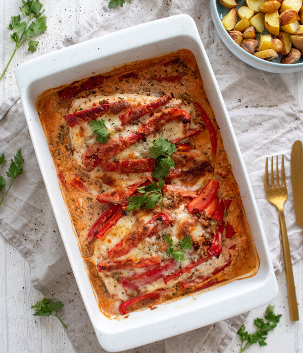 Auflaufform mit drei überbackenen Paprika-Rahmschnitzeln, gratiniert mit cremiger Soße und roten Paprikastreifen, garniert mit frischer Petersilie. Im Hintergrund goldgelbe Kartoffelwürfel auf einem Teller sowie goldenes Besteck auf einem hellen Stofftuch.