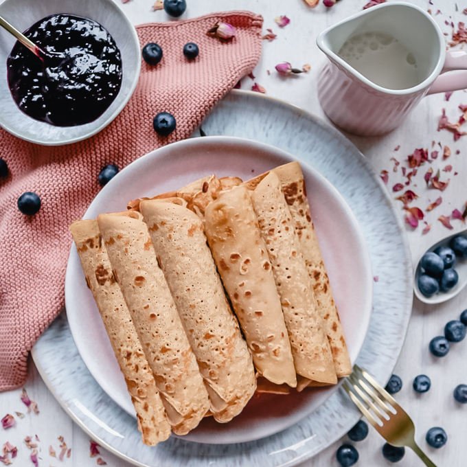 Serviervorschlag für die „RatzFatz“ Bio-Pfannkuchenmischung: Auf einem rosa Teller liegen fünf goldbraune, aufgerollte Pfannkuchen. Daneben frische Blaubeeren, ein Schälchen mit dunkler Fruchtsoße und ein Krug mit Pflanzendrink auf hellem Untergrund mit Dekor aus Blütenblättern.