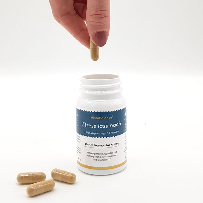Offene Kunststoffdose mit blau-weißem Etikett „Stress lass nach“ von HistaBalance®. Eine Hand hält eine braune Kapsel über die Dose, daneben drei identische Kapseln auf weißem Untergrund.