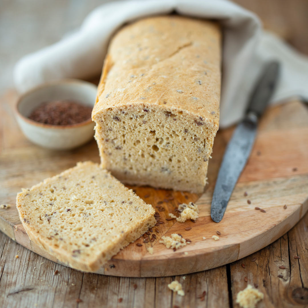 Auf einem runden Holzbrett liegt ein helles, glutenfreies Brot mit lockerer Krume und sichtbaren Saaten. Zwei Scheiben sind abgeschnitten, daneben liegen ein Messer und einige Brösel. Im Hintergrund eine Schale mit Leinsamen und ein weißes Tuch.
