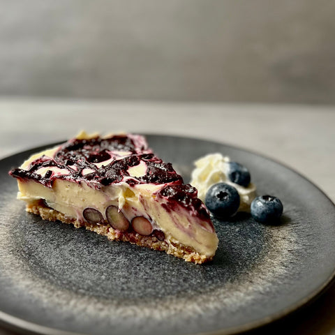 Ein Stück Heidelbeer-Cheesecake mit sichtbaren Blaubeeren im Boden, angerichtet auf einem schwarzen Teller mit Sahne und frischen Blaubeeren. 