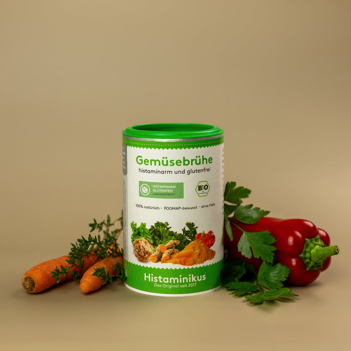 Dose „Gemüsebrühe – histaminarm und glutenfrei“ mit grünem Deckel auf beigem Hintergrund. Umgeben von frischem Gemüse: Karotten mit Grün, Petersilienblätter und rote Paprika. Aufdruck mit Zutatenabbildung und Bio-Siegel.
