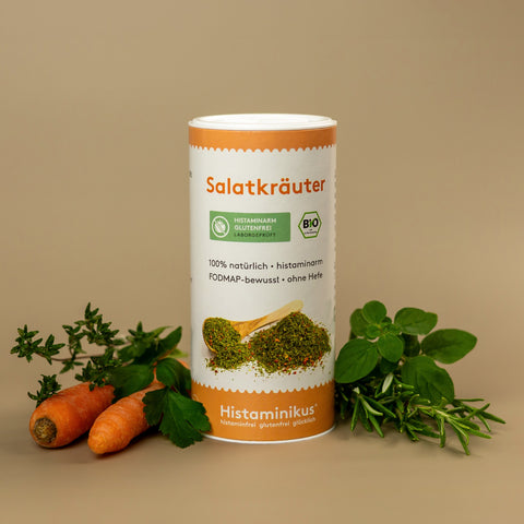 Foto der Gewürzdose „Salatkräuter“ von Histaminikus mit natürlicher Dekoration. Auf dem Etikett stehen Hinweise wie „100 % natürlich“, „histaminarm“, „FODMAP-bewusst“ und „ohne Hefe“. Unten Markenlogo mit Claim. Im Vordergrund frische Karotten, Kräuter und grüne Blätter.
