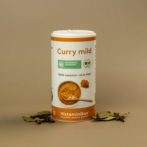 Dose „Curry mild“ mit orange-weißem Etikett auf beigem Hintergrund. Darauf Bio- und histaminarm-Siegel, Text: 100 % natürlich, ohne Hefe. Umgeben von Lorbeerblättern und Gewürznelken.