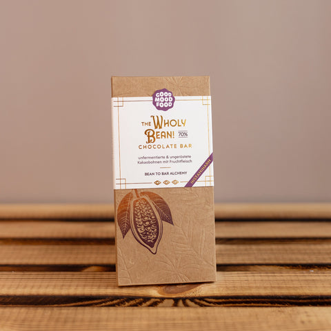 Verpackte Schokolade von Good Mood Food mit der Aufschrift ‚The Wholy Bean! 70 % Chocolate Bar‘ auf einem Holztisch. Die Verpackung ist braun mit weißem Etikett und zeigt eine stilisierte Kakaofrucht.
