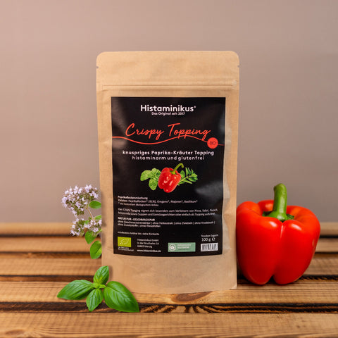 Produktfoto: Braune Standbodenverpackung „Crispy Topping“ mit schwarzem Etikett auf Holzuntergrund. Daneben frische Kräuter (Basilikum, Majoran) und eine rote Paprika. Hintergrund neutral beige.