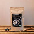 Braune Papierverpackung mit schwarzem Etikett. Aufschrift: „Histaminikus – Knusperpower Müsli“. Darunter: „Knuspermüsli mit Macadamianüssen, histaminarm und glutenfrei“. Mittig ein rundes Foto mit Müsli, Joghurt und Blaubeeren in einer Schale sowie eine kleinere Nahaufnahme des Müslis. Im Vordergrund liegen einige Blaubeeren und ein Holzlöffel mit Haferflocken auf einem Holztisch.