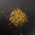 Loser Kräutertee in goldgrüner Farbgebung auf schwarzem Untergrund, Mischung mit erkennbaren Blatt-, Blüten- und Stängelanteilen, darunter feinere gelbliche und bräunliche Bestandteile.