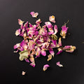 Eine Handvoll getrockneter rosa Rosenblütenblätter locker auf schwarzem Untergrund verteilt.