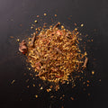 Häufchen loser Wintertee auf schwarzem Untergrund. Die Mischung enthält erkennbar getrocknete Blüten, Hibiskusstücke, Zimtrinde, ganze Nelken und weitere pflanzliche Bestandteile in rot-braunen und goldgelben Tönen. Einzelne Krümel und Fasern sind rund um den Haufen verstreut.