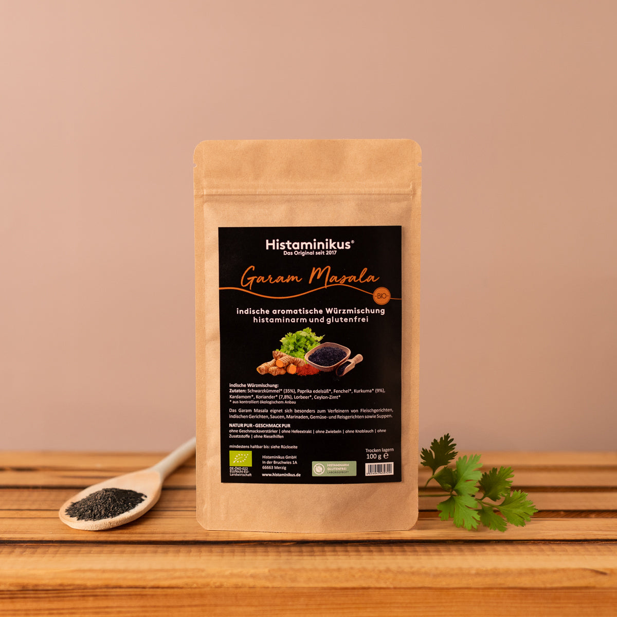 Braune Packung „Garam Masala“ von Histaminikus steht auf einem Holzuntergrund vor hellbraunem Hintergrund. Daneben ein Karianderzweig und ein Holzlöffel mit Kümmel. Die Würzmischung ist histaminarm, glutenfrei und bio-zertifiziert.