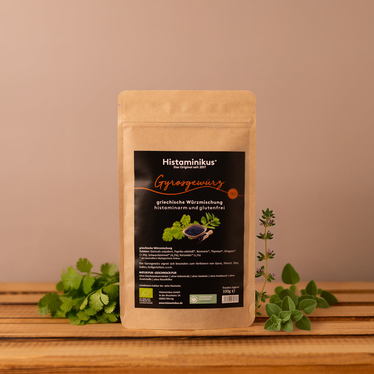 Braune Gewürztüte mit schwarzem Etikett der Marke Histaminikus. Aufschrift: „Gyrosgewürz – griechische Würzmischung, histaminarm und glutenfrei“. Darunter Abbildung von Gewürzen und Kräutern. Die Tüte steht auf einem Holzbrett, dekoriert mit frischen Kräutern vor beigem Hintergrund.