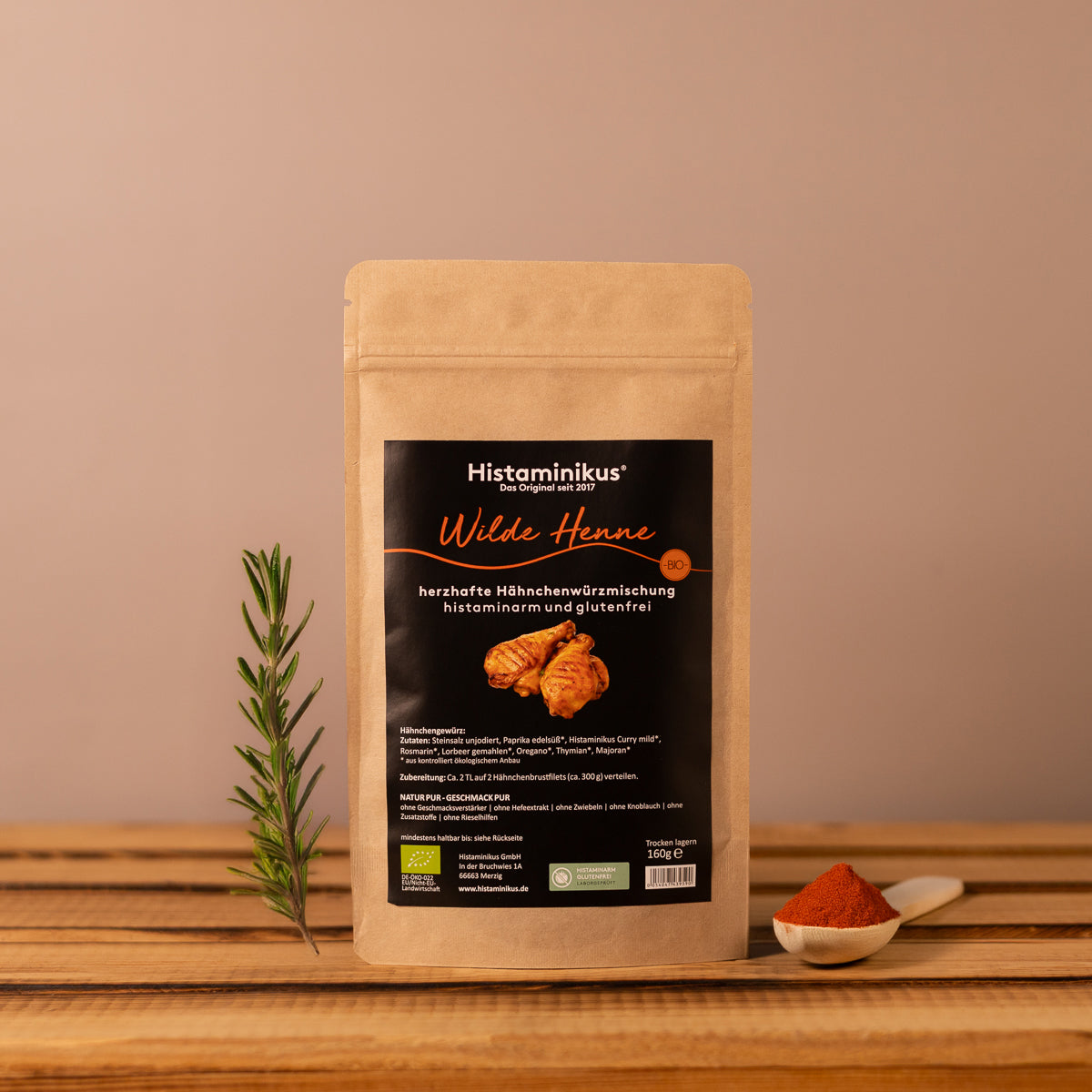 Braune Gewürztüte mit schwarzem Etikett der Marke Histaminikus. Aufschrift: „Wilde Henne – herzhafte Hähnchenwürzmischung, histaminarm und glutenfrei“. Darunter Abbildung von gewürzten Hähnchenteilen. Die Tüte steht auf einem Holzbrett, dekoriert mit Rosmarinzweig und einem Löffel mit Paprikapulver vor beigem Hintergrund.