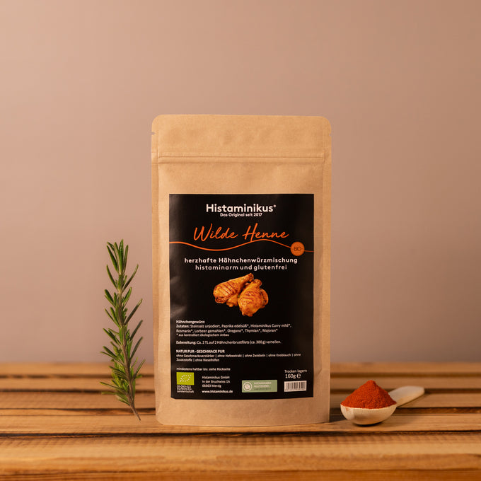Braune Gewürztüte mit schwarzem Etikett der Marke Histaminikus. Aufschrift: „Wilde Henne – herzhafte Hähnchenwürzmischung, histaminarm und glutenfrei“. Darunter Abbildung von gewürzten Hähnchenteilen. Die Tüte steht auf einem Holzbrett, dekoriert mit Rosmarinzweig und einem Löffel mit Paprikapulver vor beigem Hintergrund.