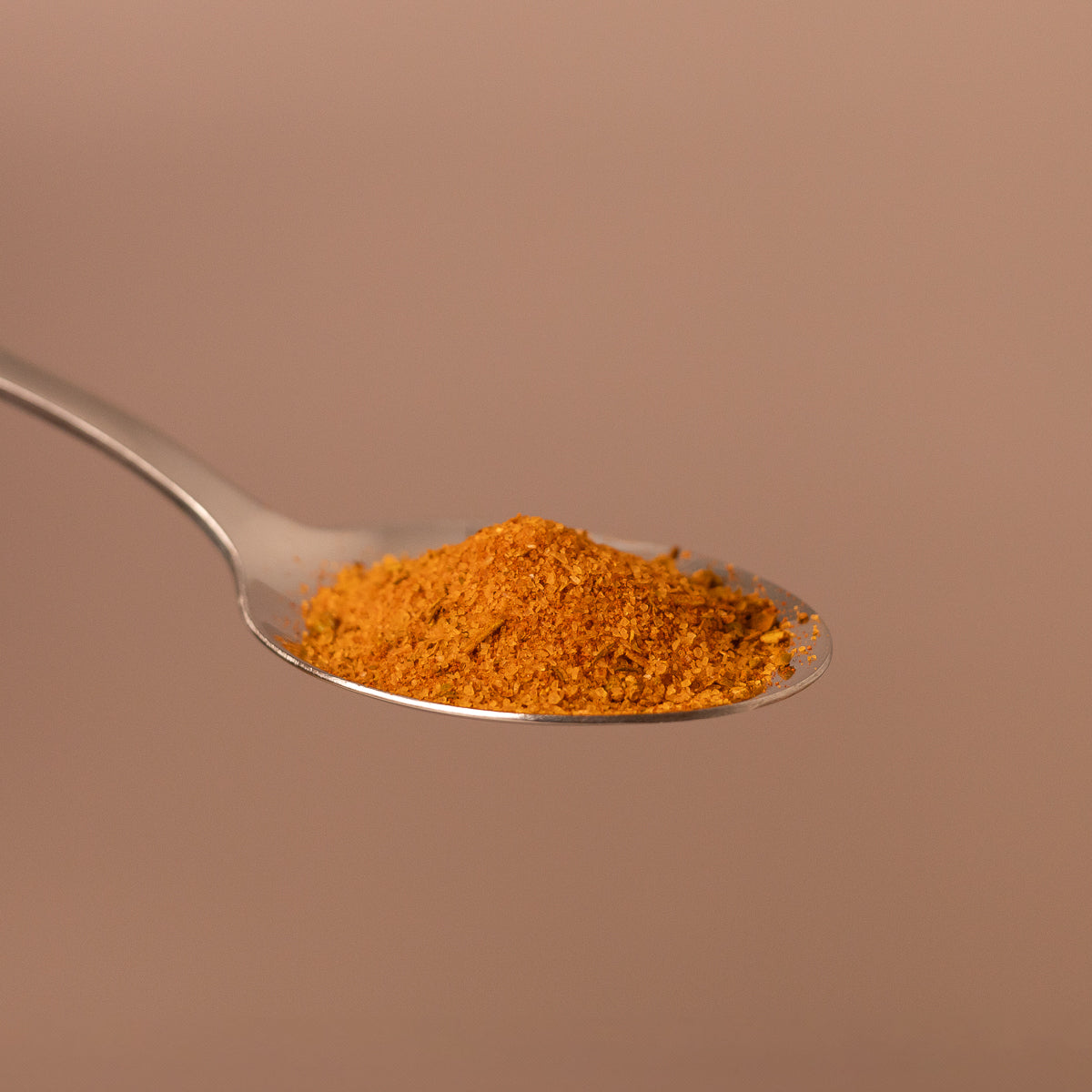 Silberner Löffel mit einem gehäuften Haufen fein gemahlener, orange-brauner Gewürzmischung vor einfarbigem Hintergrund.