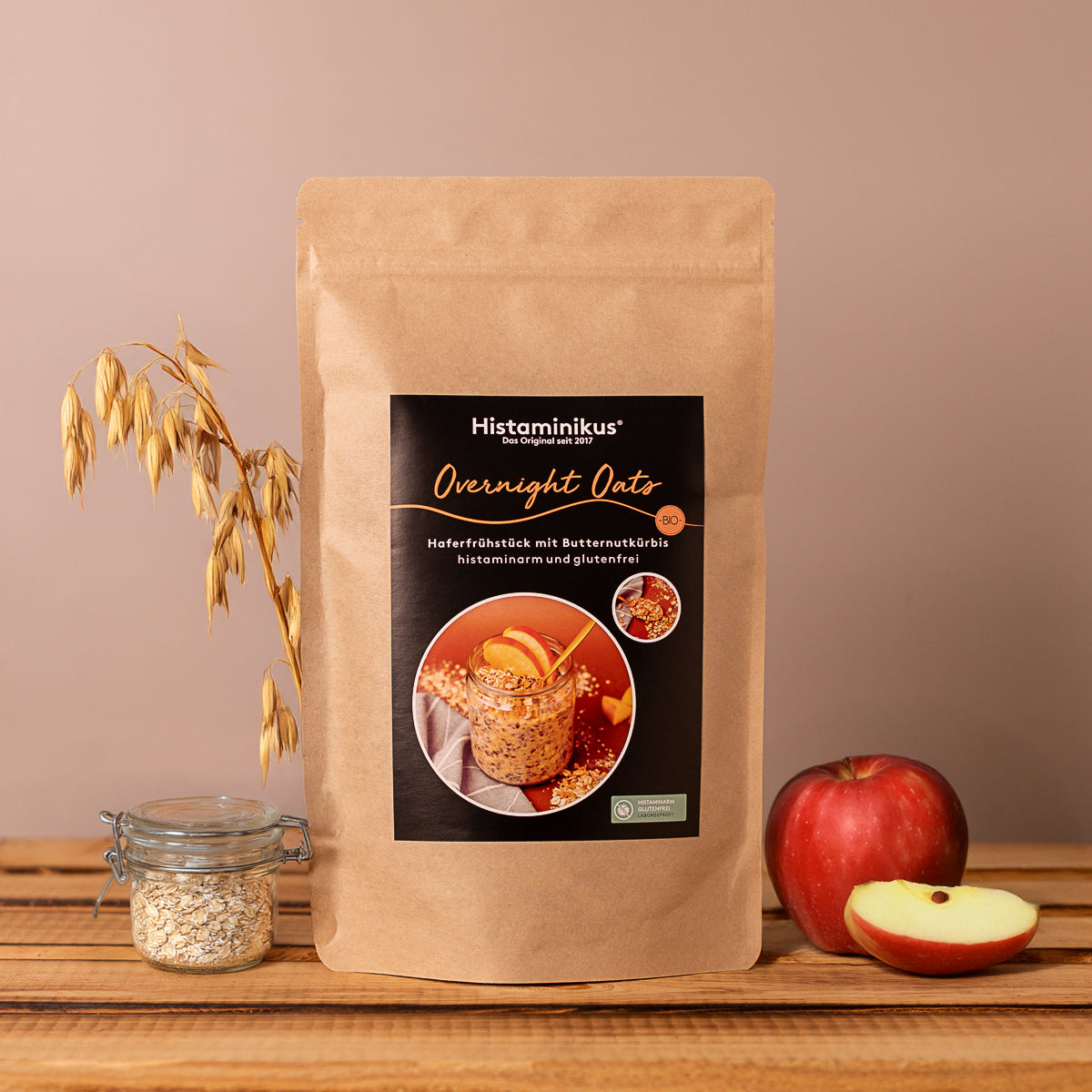 Vorderansicht einer braunen, stehenden Papiertüte mit dem Produktetikett „Histaminikus Overnight Oats“. Auf dem Etikett sind ein Glas mit Haferfrühstück, Apfelscheiben und Kürbisstückchen sowie der Text „Haferfrühstück mit Butternutkürbis, histaminarm und glutenfrei“ zu sehen. Links neben der Tüte stehen ein Glas mit Haferflocken und ein Bündel Haferhalme, rechts ein roter Apfel mit einer angeschnittenen Apfelscheibe. Der Hintergrund ist beige, der Untergrund eine Holzfläche.