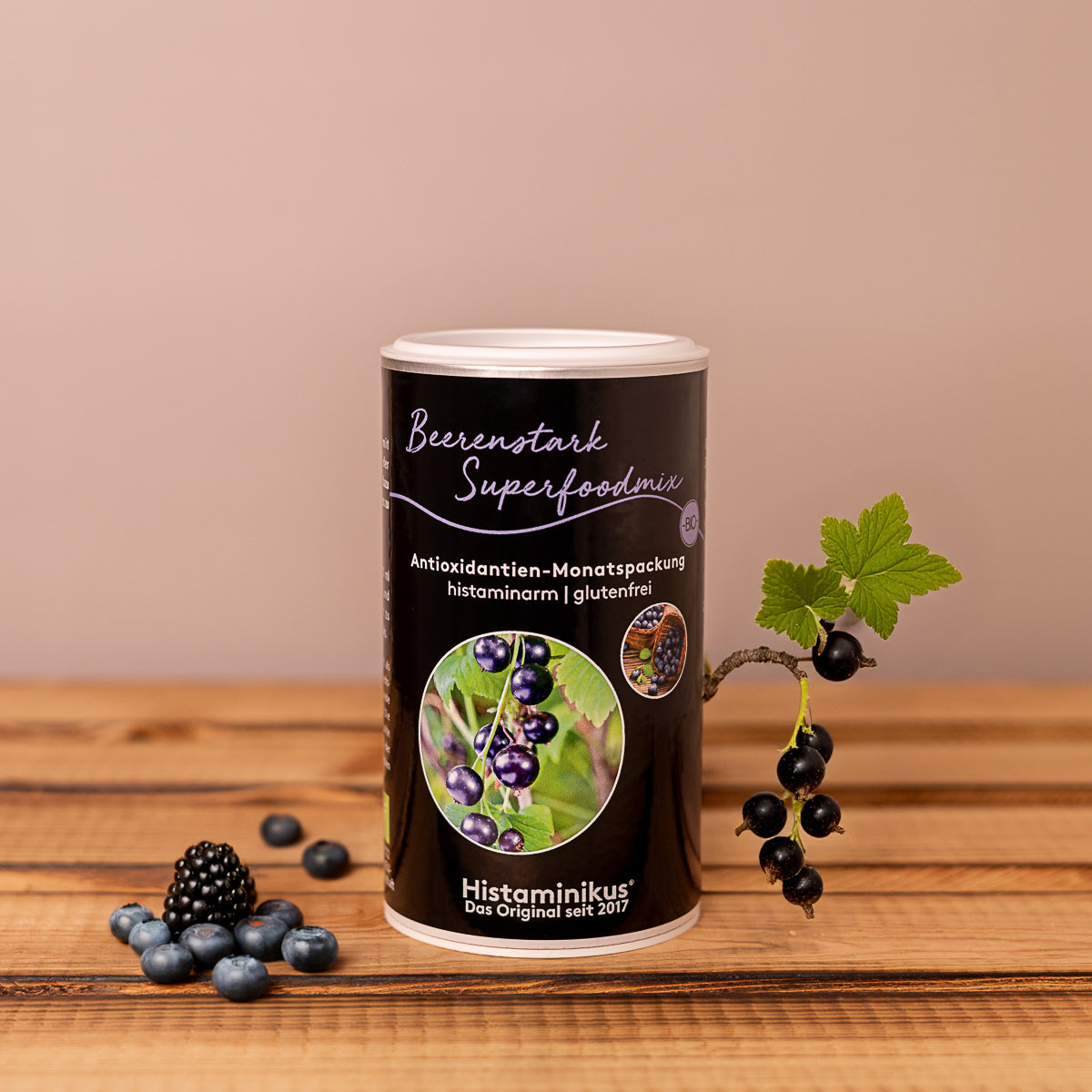 Dose mit dem Etikett „Beerenstark Superfoodmix“ von Histaminikus, stehend auf einer Holzoberfläche vor beigem Hintergrund. Die Dose zeigt Bilder von schwarzen Johannisbeeren und trägt den Text „Antioxidantien-Monatspackung, histaminarm, glutenfrei“. Vor der Dose liegen eine Brombeere und mehrere Heidelbeeren, daneben ein Zweig mit schwarzen Johannisbeeren und einem grünen Blatt.