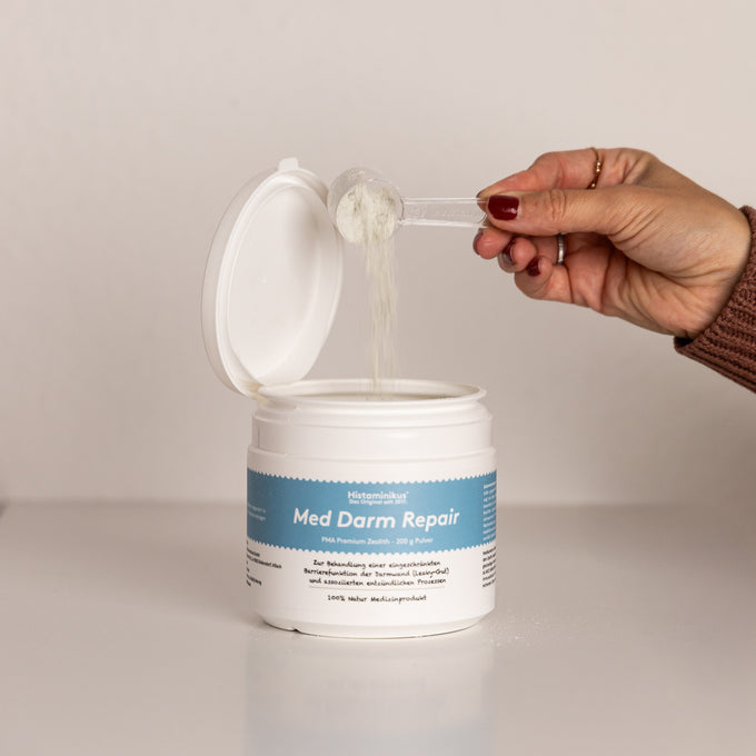 Hand mit rot lackierten Fingern hält einen Messlöffel über eine geöffnete Dose „Med Darm Repair“, während feines Pulver in die Dose fällt.