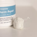 Nahaufnahme eine weißen Dose mit blauem Etikett „Med Darm Repair“ und einem transparenten Messlöffel, gefüllt mit feinem, hellgrauem Zeolithpulver.