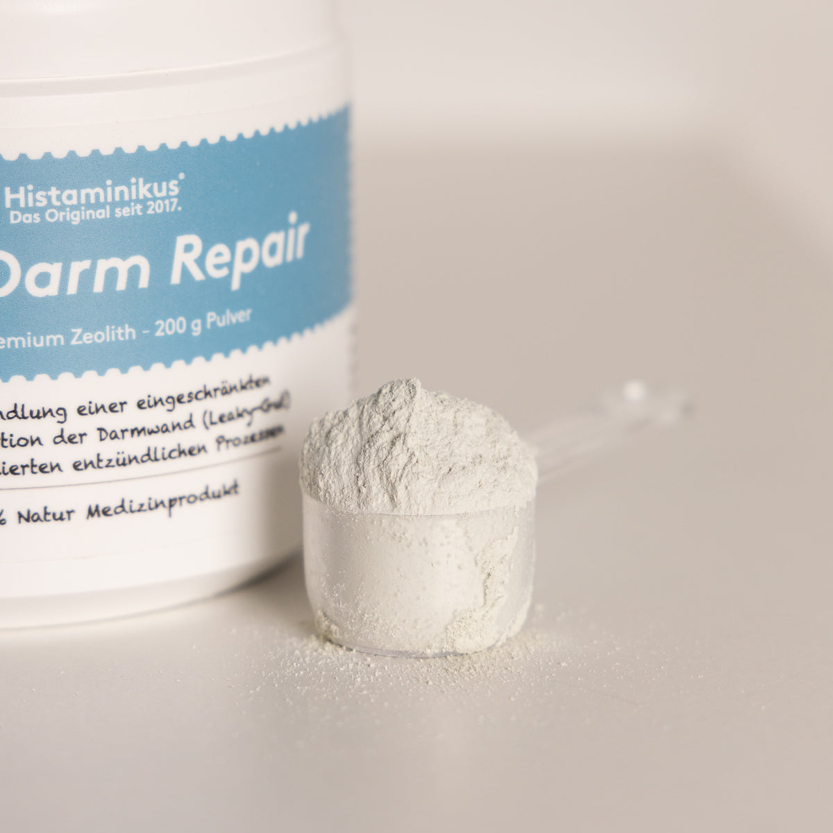 Nahaufnahme eine weißen Dose mit blauem Etikett „Med Darm Repair“ und einem transparenten Messlöffel, gefüllt mit feinem, hellgrauem Zeolithpulver.