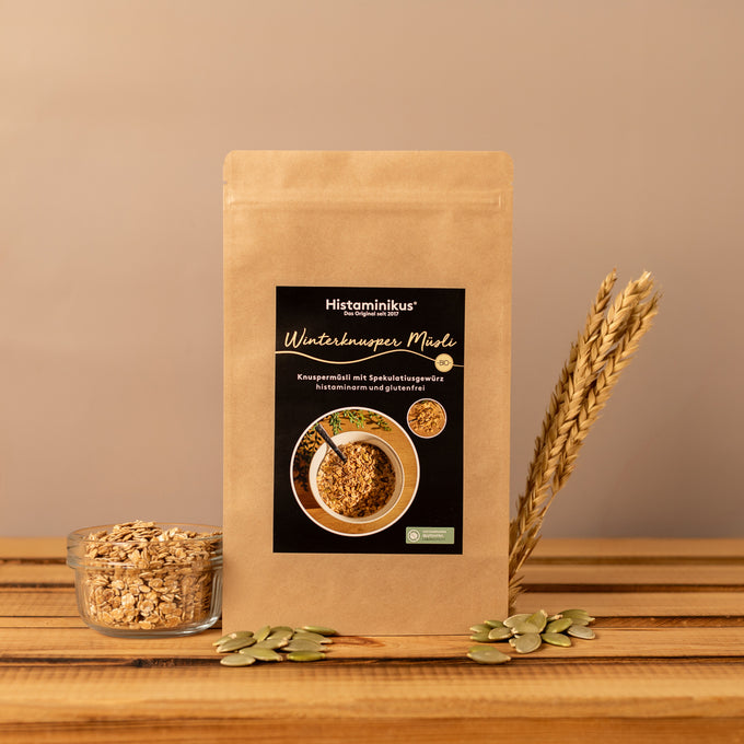 Stehende Papierverpackung „Winterknusper Müsli“ von Histaminikus auf einer Holzoberfläche, davor eine Glasschale mit Haferflocken und verstreute Kürbiskerne, rechts daneben Getreideähren, warmes beigefarbenes Hintergrundlicht.