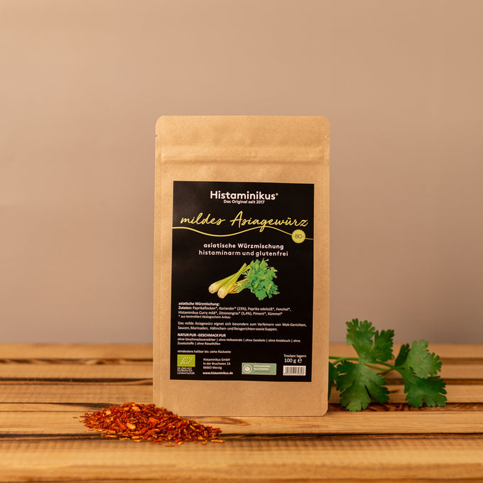 Stehende Papierverpackung „Mildes Asiagewürz“ von Histaminikus auf einer Holzoberfläche, davor ein kleines Häufchen roter Paprikaflocken, rechts frischer Koriander, neutraler beigefarbener Hintergrund.
