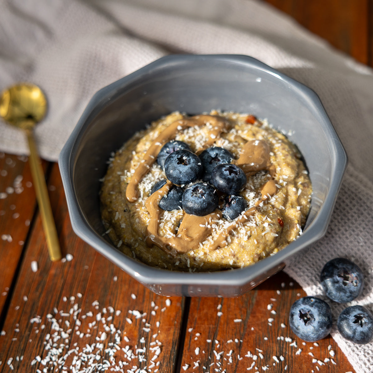 Graue Schale mit cremigem Polentafrühstück auf einem dunklen Holztisch. Das Frühstück ist mit Heidelbeeren, einem Topping aus Nussmus und feinen Kokosraspeln garniert. Im Hintergrund liegt ein helles Tuch, links daneben ein goldener Löffel, auf dem Tisch sind zusätzlich Kokosraspeln und einzelne Heidelbeeren verteilt.