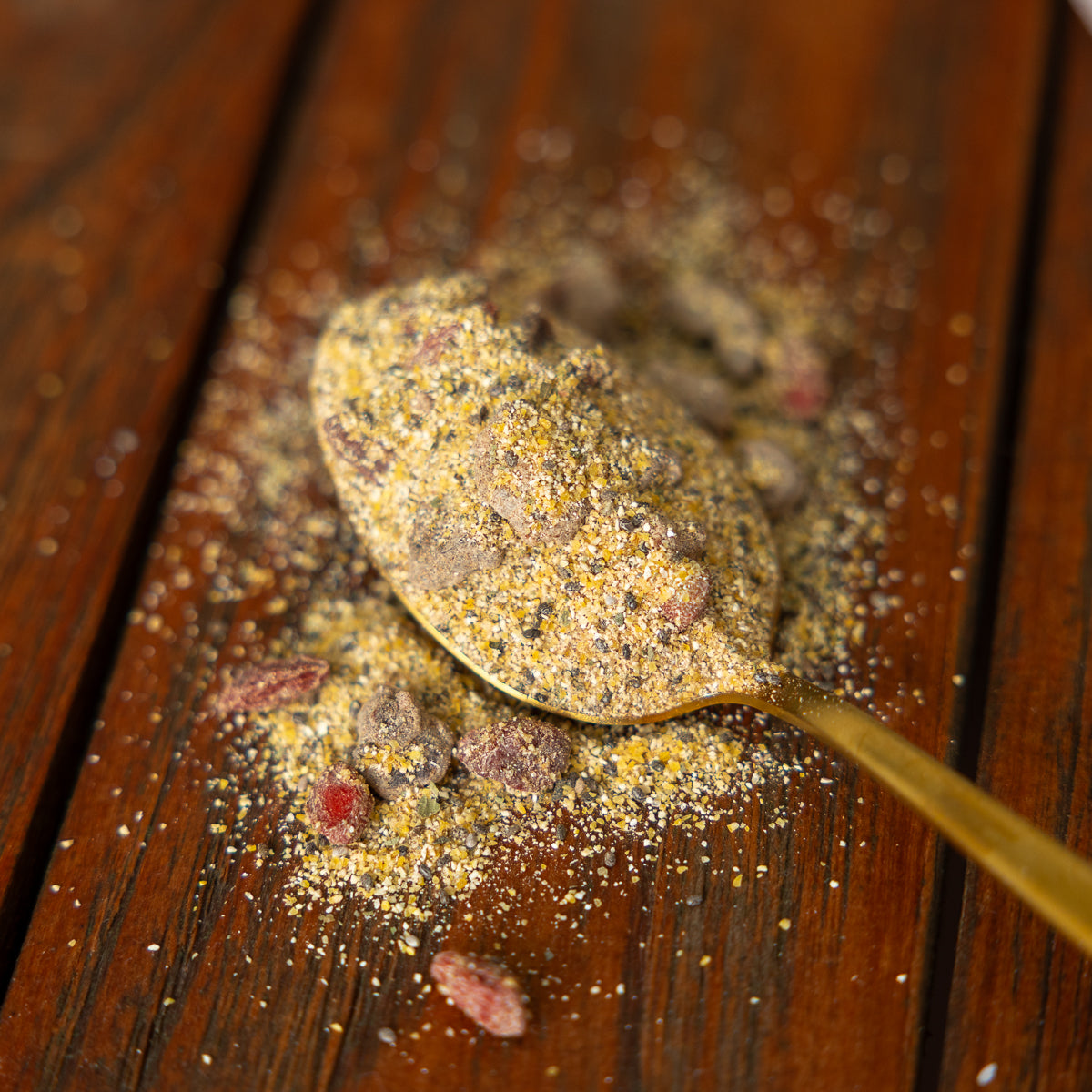 Goldener Löffel mit einer trockenen Polentamischung auf einem dunklen Holztisch. In der Mischung sind feine Körner sowie sichtbare Stückchen, darunter Gojibeeren, enthalten. Rund um den Löffel liegt lose Mischung verstreut auf der Holzoberfläche.