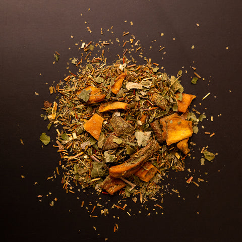 Teemischung aus Rooibos mit getrockneten Mangostücken und Kräutern, lose auf dunklem Hintergrund ausgebreitet.