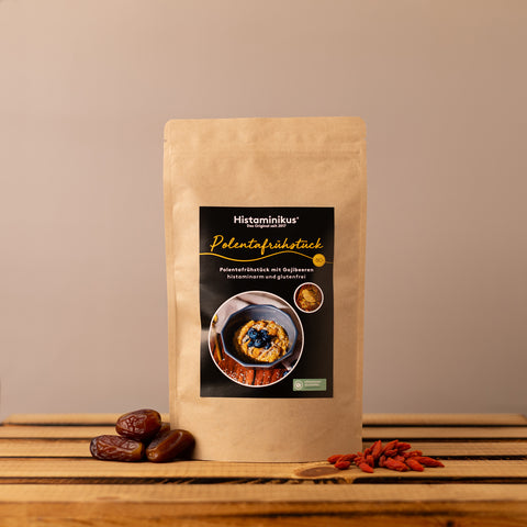 Standbodenbeutel aus braunem Kraftpapier mit schwarzem Etikett „Polentafrühstück“. Auf dem Etikett steht „Polentafrühstück mit Gojibeeren, histaminarm und glutenfrei“, dazu ist eine Schale mit Polenta und dunklen Beeren abgebildet. Der Beutel steht auf einem rustikalen Holztisch vor beigefarbenem Hintergrund, links liegen Datteln, rechts Gojibeeren.