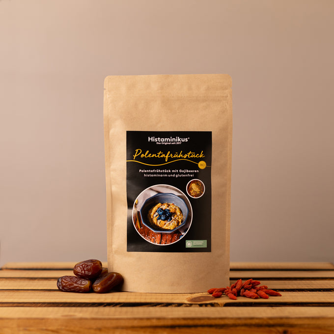 Standbodenbeutel aus braunem Kraftpapier mit schwarzem Etikett „Polentafrühstück“. Auf dem Etikett steht „Polentafrühstück mit Gojibeeren, histaminarm und glutenfrei“, dazu ist eine Schale mit Polenta und dunklen Beeren abgebildet. Der Beutel steht auf einem rustikalen Holztisch vor beigefarbenem Hintergrund, links liegen Datteln, rechts Gojibeeren.