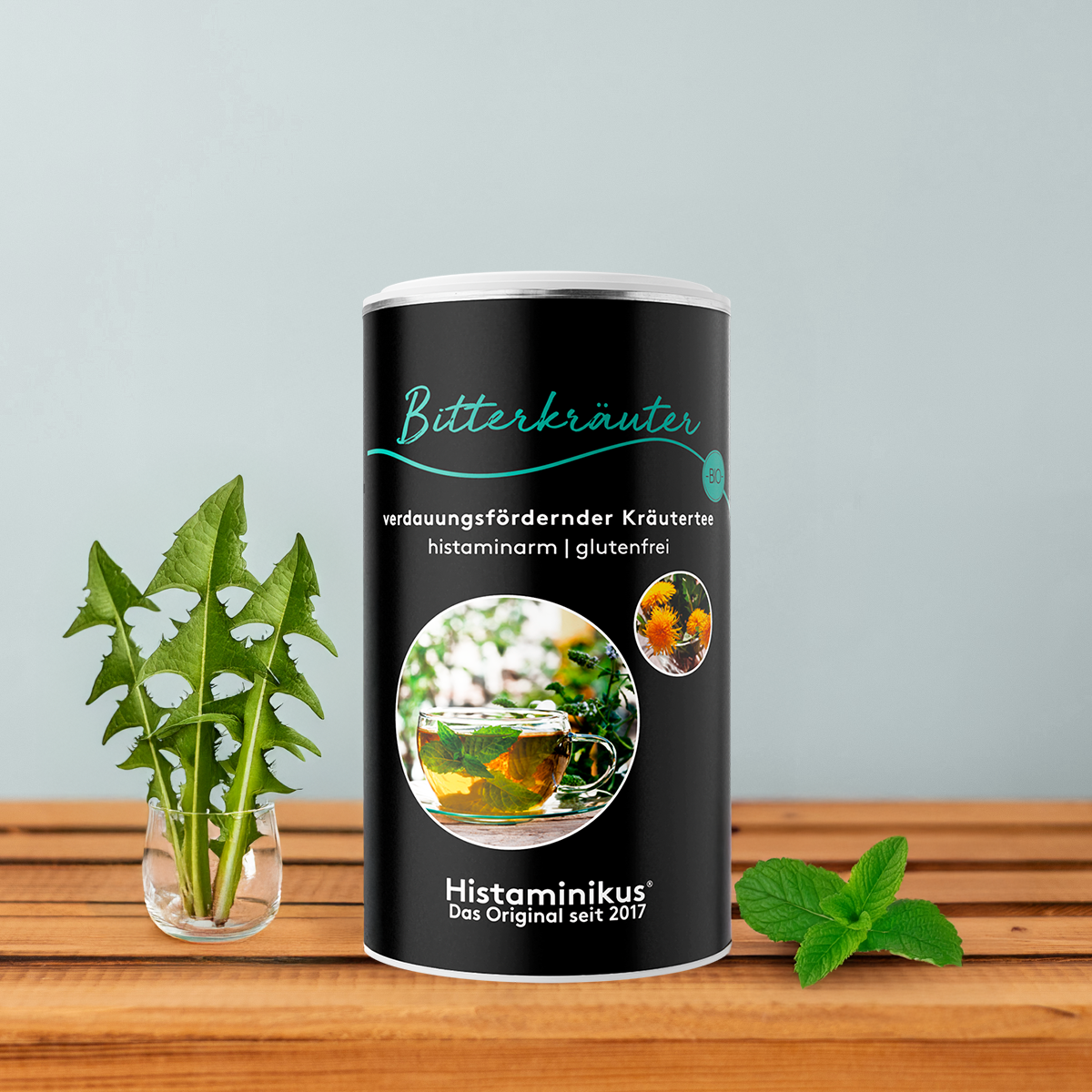 Schwarze Teedose „Bitterkräuter“ mit türkisfarbener Schrift und Abbildung einer Teetasse, dekoriert mit Löwenzahnblättern und Minze vor hellgrauem Hintergrund. 