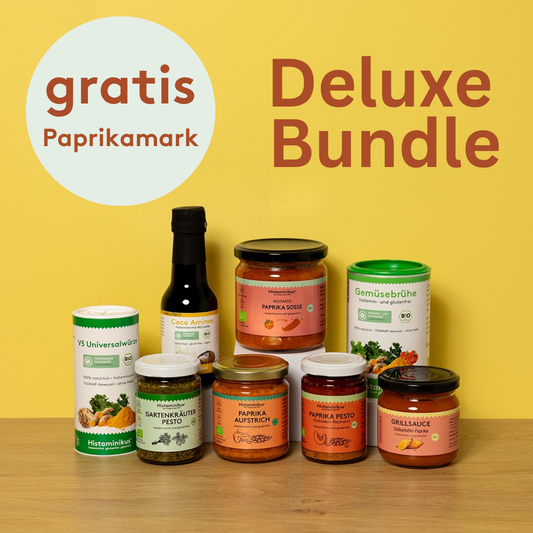 Acht histaminarme Bio-Produkte von Histaminikus auf hellem Holztisch vor gelbem Hintergrund: zwei Gewürzdosen, fünf Gläser mit Paprika-Aufstrichen, -Pesto, -Mark, -Soße und Grillsauce sowie eine Flasche Coco Aminos. Darüber ein Kreis mit rotem Schriftzug „gratis Paprikamark“.