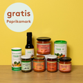 Acht histaminarme Bio-Produkte von Histaminikus auf hellem Holztisch vor gelbem Hintergrund: zwei Gewürzdosen, fünf Gläser mit Paprika-Aufstrichen, -Pesto, -Mark, -Soße und Grillsauce sowie eine Flasche Coco Aminos. Darüber ein Kreis mit rotem Schriftzug „gratis Paprikamark“.