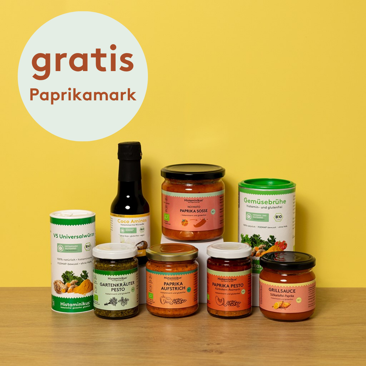 Acht histaminarme Bio-Produkte von Histaminikus auf hellem Holztisch vor gelbem Hintergrund: zwei Gewürzdosen, fünf Gläser mit Paprika-Aufstrichen, -Pesto, -Mark, -Soße und Grillsauce sowie eine Flasche Coco Aminos. Darüber ein Kreis mit rotem Schriftzug „gratis Paprikamark“.