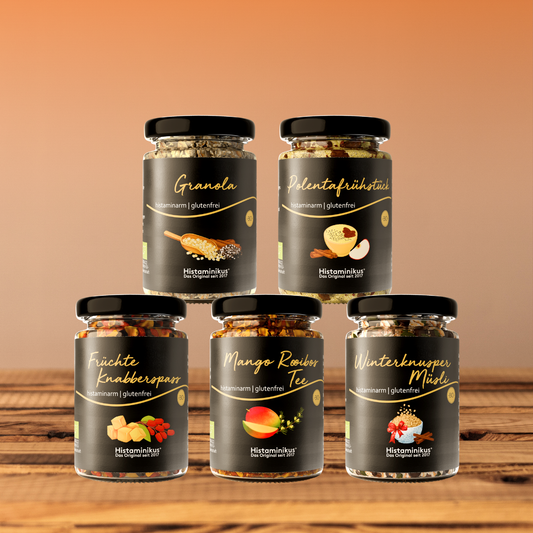 Fünf histaminarme, glutenfreie Frühstücksprodukte von Histaminikus in Gläsern: Granola, Polentafrühstück, Früchte Knabberspaß, Mango Rooibos Tee und Winterknusper Müsli, arrangiert auf einem Holztisch vor warmem Farbverlauf-Hintergrund.