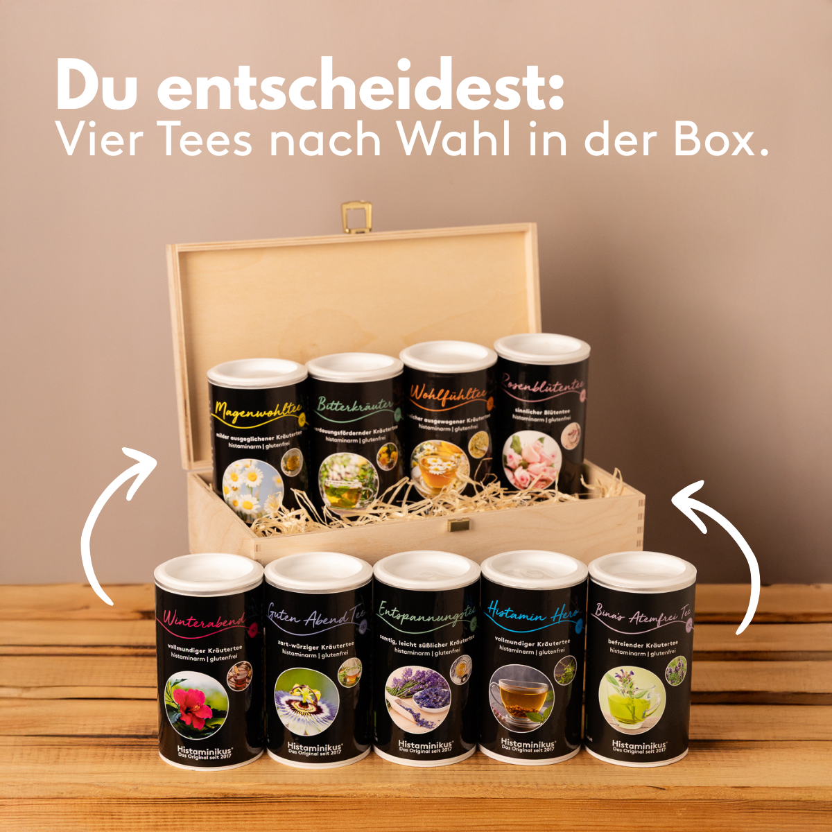 Neun verschiedene Teesorten in schwarzen Dosen stehen vor und in einer geöffneten Holzbox auf einem Holztisch. Darüber steht der Schriftzug: „Du entscheidest: Vier Tees nach Wahl in der Box.“ Zwei weiße Pfeile zeigen auf die Box mit den Teedosen.